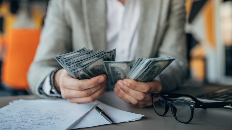 A person in a suit counts money by hand at a desk with papers, a pen, and glasses, showing how to count money accurately