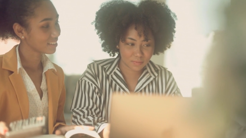 Two women sit together looking at a laptop screen while discussing financial information