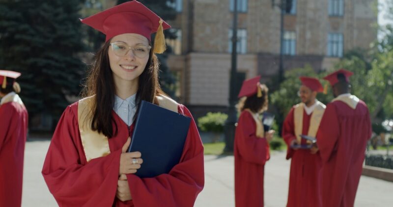 A young woman in a red graduation gown and cap smiles while holding a diploma folder, standing outdoors on a university campus