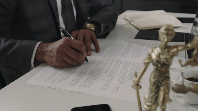 A professional in a dark suit writing on a legal document at a desk, with a golden Lady Justice statue in the foreground