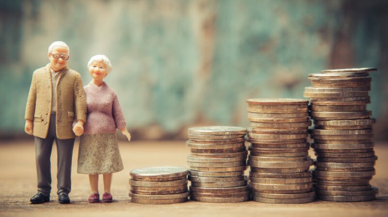 Miniature elderly couple standing next to stacks of coins symbolizing retirement savings