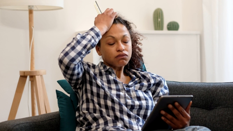 A worried woman sits on a couch holding a tablet and thinking about her payday loan debt problems