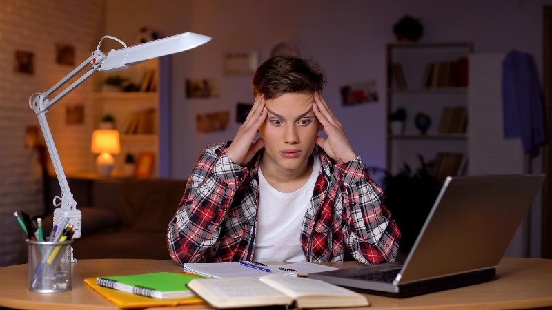 A stressed student sitting at a desk with a laptop and study materials while preparing for an oral and written math test