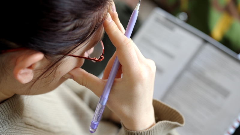 A student holding a pen and reviewing notes with a focused, slightly frustrated expression during written math test preparation
