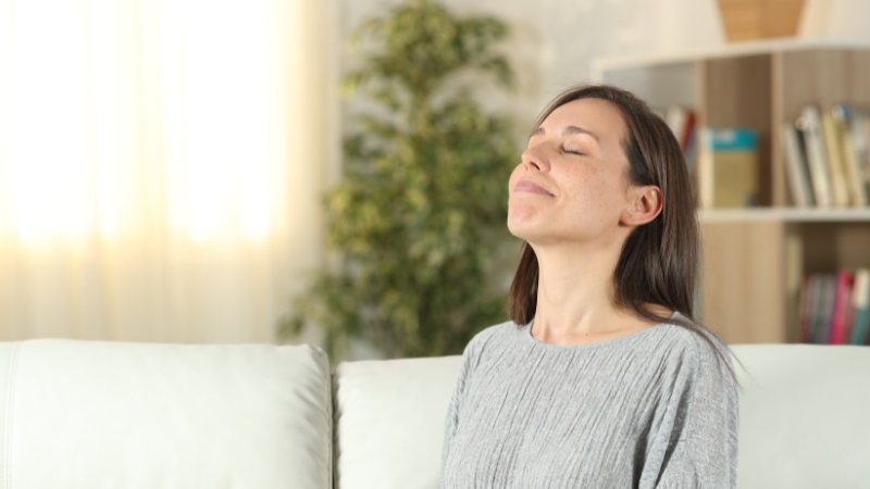 A woman sits calmly on a sofa with her eyes closed, practicing a one minute breathing reset to ease anxiety