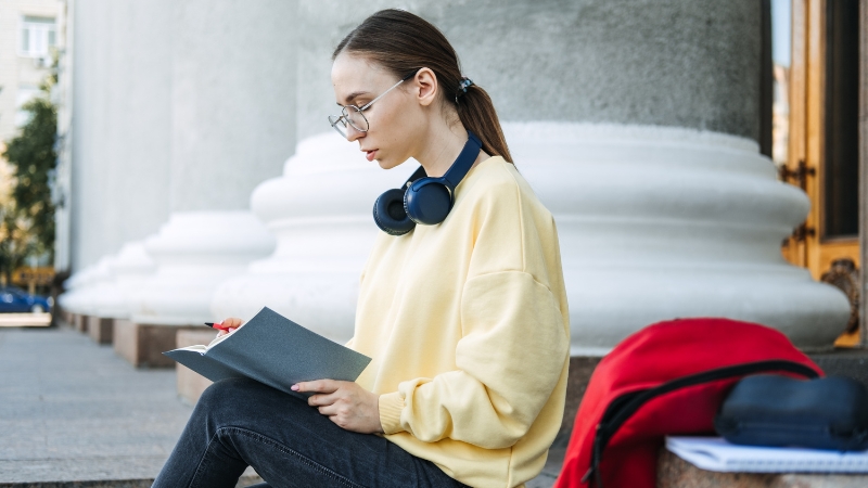 A young woman sits outside writing in a notebook, taking a moment for a positive affirmation to start her day calmly