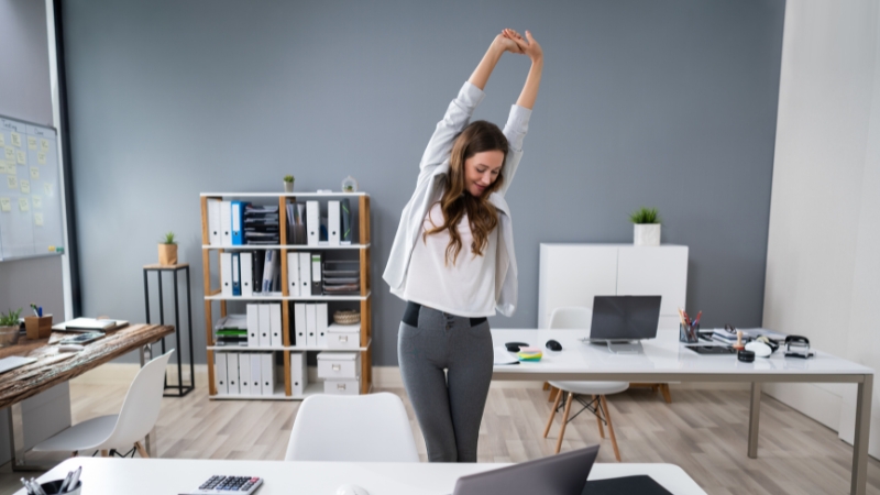 A woman stands in an office stretching her arms overhead to release tension and feel calmer