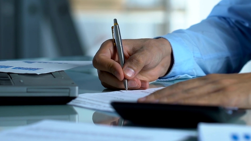 A person in a blue shirt reviews documents and takes notes with a pen at a desk