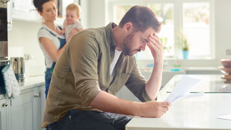 A man looks stressed while reading a bill at the kitchen counter as a woman holds a baby in the background